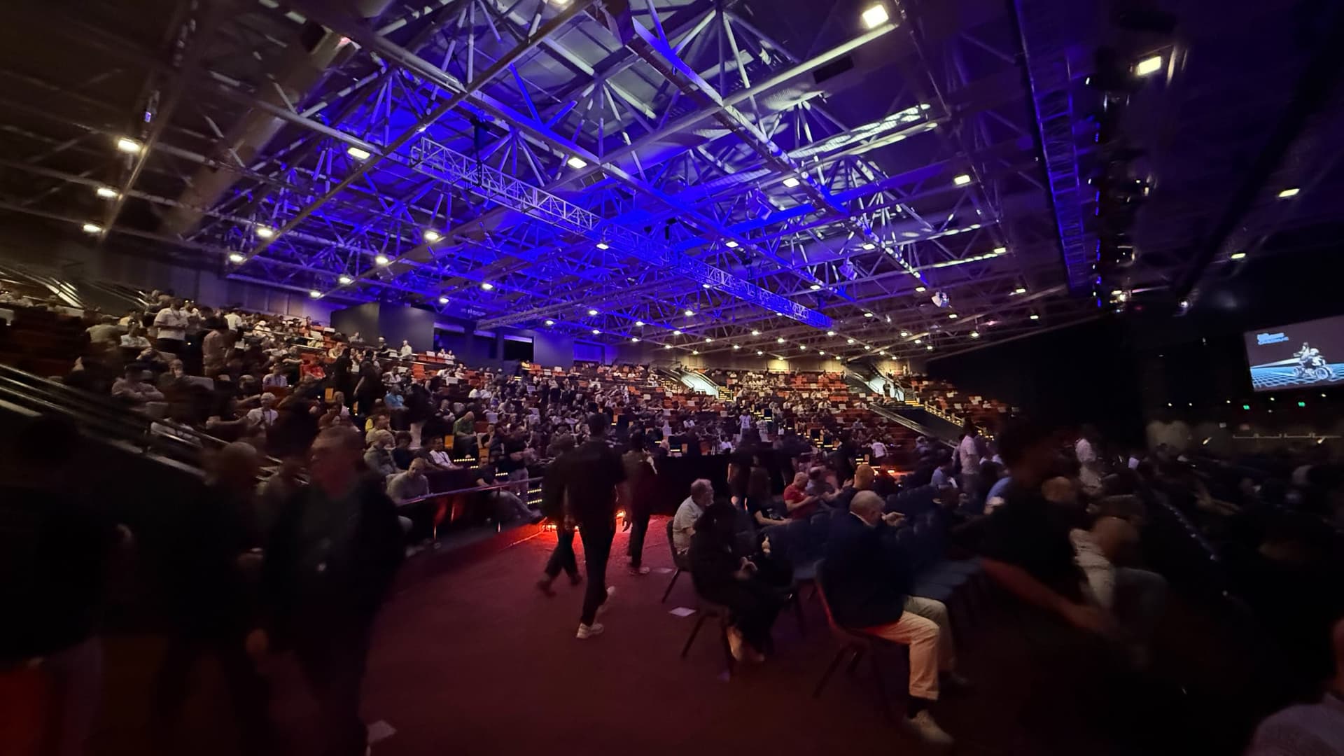 A packed auditorium under blue-lit roof trusses at BSides Canberra 2026